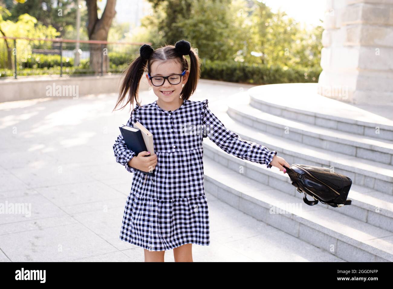 Joli petit enfant fille 5-6 ans jouer à l'extérieur porter des lunettes,  robe élégante tenant sac à dos. Retour à l'école. Un joli pupille souriant  se prépare pour f Photo Stock -