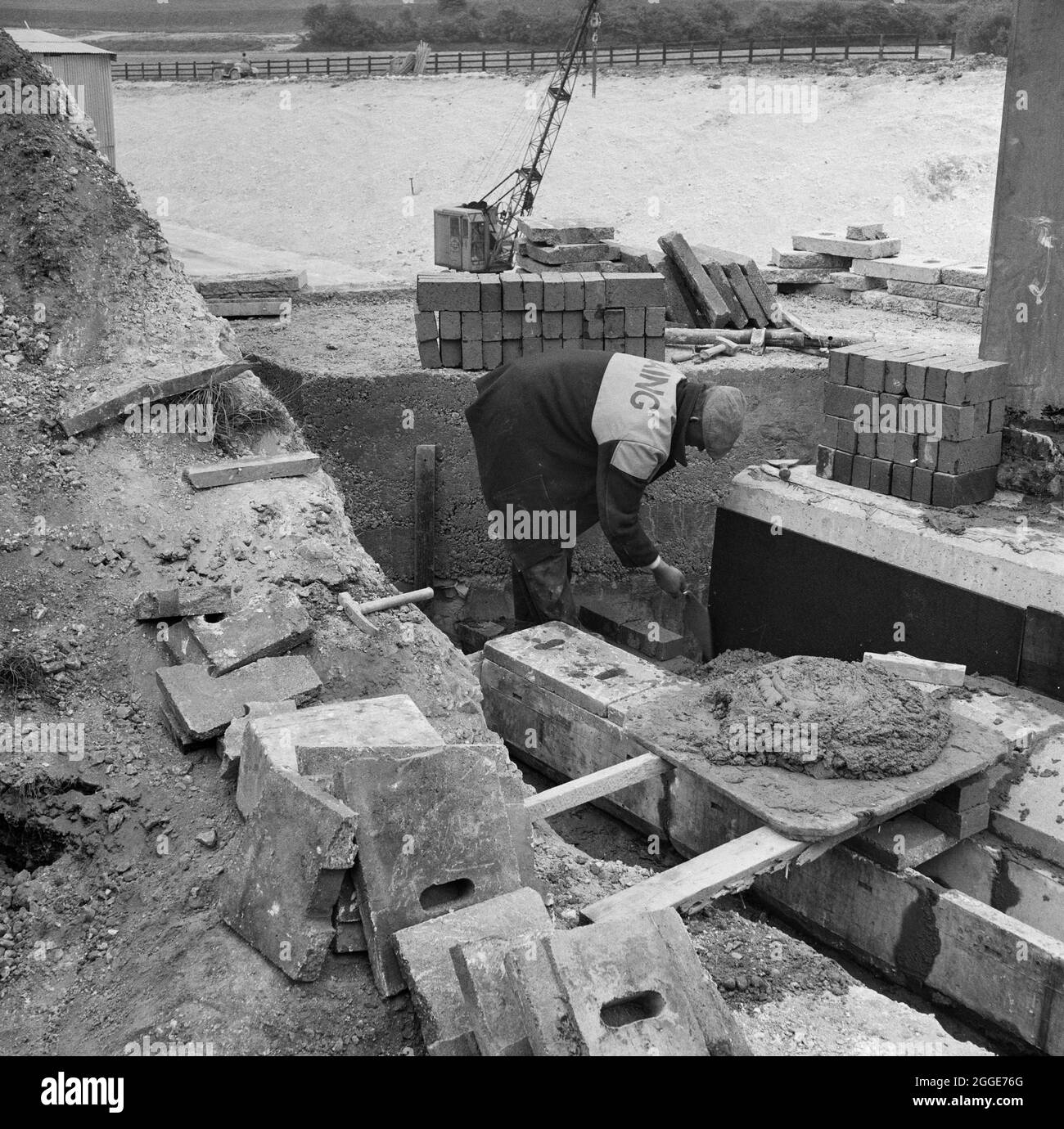 Un ouvrier de Laing posant des briques près d'un canal de drainage pendant la construction de la structure 9, un pont transportant la ligne de chemin de fer Luton-Dunstable au-dessus de l'autoroute de Londres à Yorkshire (M1). La légende sous l'impression de l'album correspondant indique qu'il s'agit de « brickwork bleu ». Banque D'Images