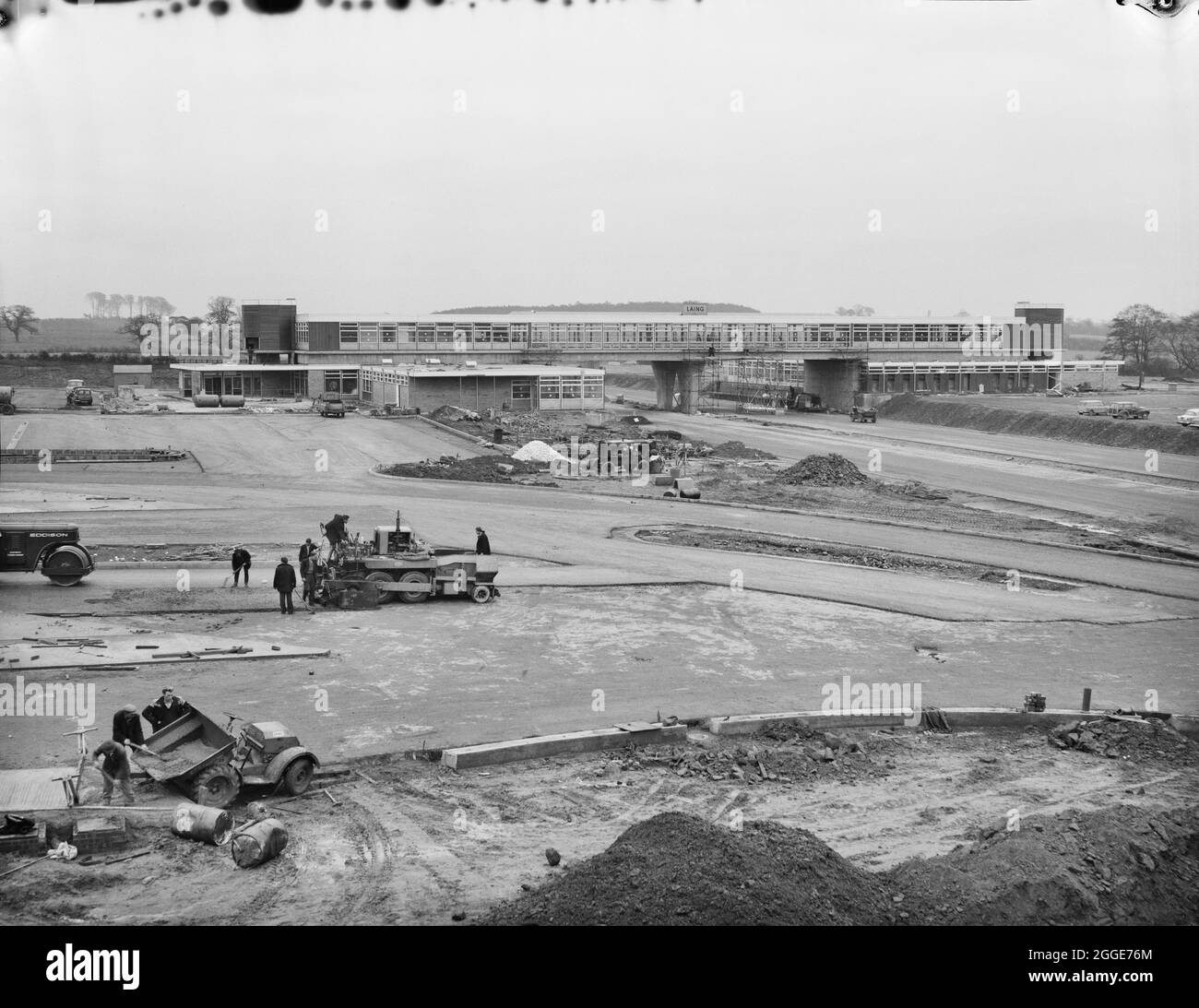 Vue sur la zone de service de Keele pendant la construction de l'autoroute Birmingham-Preston (M6), montrant une équipe de travailleurs dans le tarmac de premier plan. Dans l'album, la légende dactylographiée ci-dessous la photo se lit "ce développement réalisé par la Société pour les services d'autoroute limitée se compose du restaurant principal au-dessus des cafétérias de l'autoroute et des cafés de transport des deux côtés de l'autoroute. Il s'agit d'une version beaucoup améliorée de celle de Newport Pagnell. » Banque D'Images