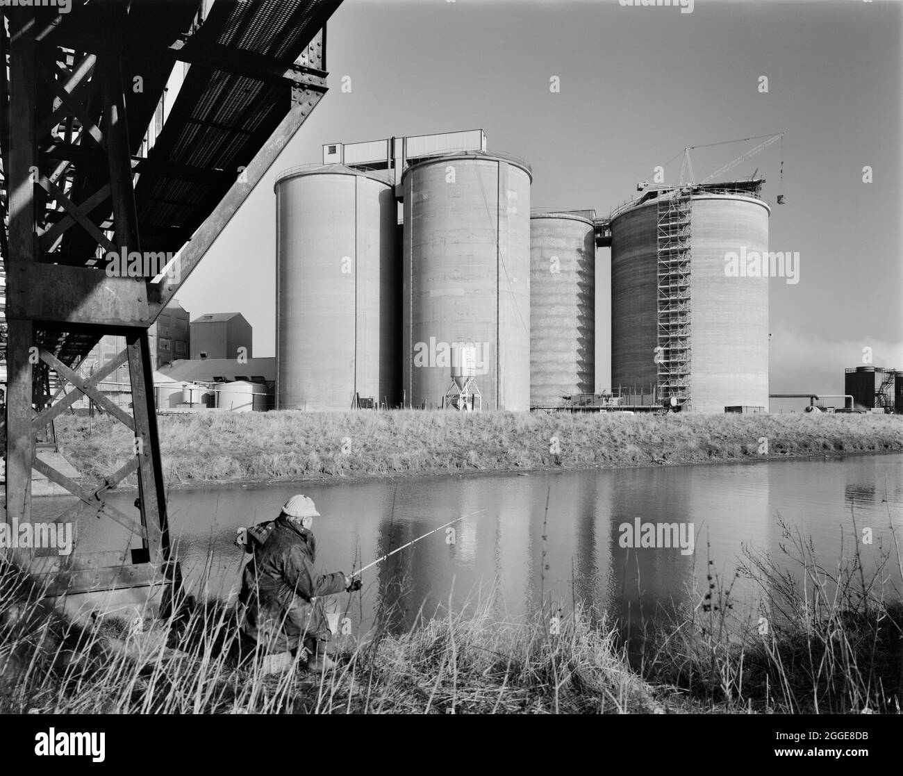 Vue sur un homme pêchant sur la rivière Witham vers quatre silos à sucre à la fabrique de sucre de Bardney, montrant la construction de l'un d'eux en voie d'achèvement. Cette image a été cataloguée dans le cadre du projet Breaking New Ground en partenariat avec la John Laing Charitable Trust en 2019-20. Banque D'Images