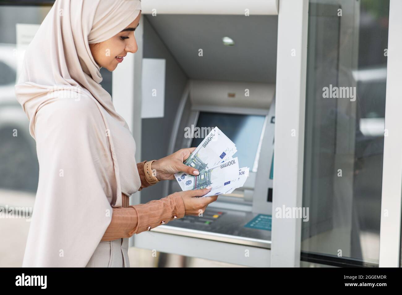 Bonne jolie femme islamique millénaire dans le hijab comptant beaucoup d'euros près de atm extérieur. Transfert de fonds, compte financier personnel, réception d'argent, W Banque D'Images