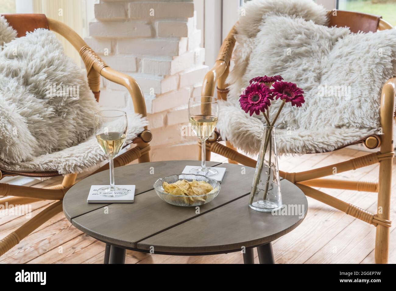 Ambiance chaleureuse en fin d'après-midi : deux chaises en osier avec oreillers et une table basse avec des verres à vin et un en-cas, préparés comme solarium dans un jardin d'hiver Banque D'Images