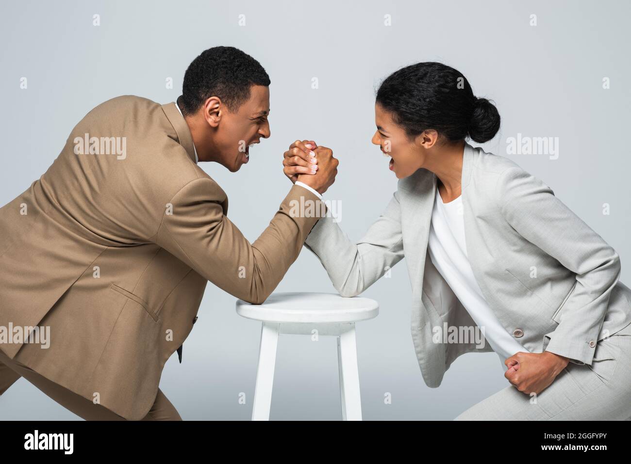 vue latérale de la femme d'affaires afro-américaine et de l'homme qui fait le bras lutte sur la chaise blanche isolée sur gris Banque D'Images