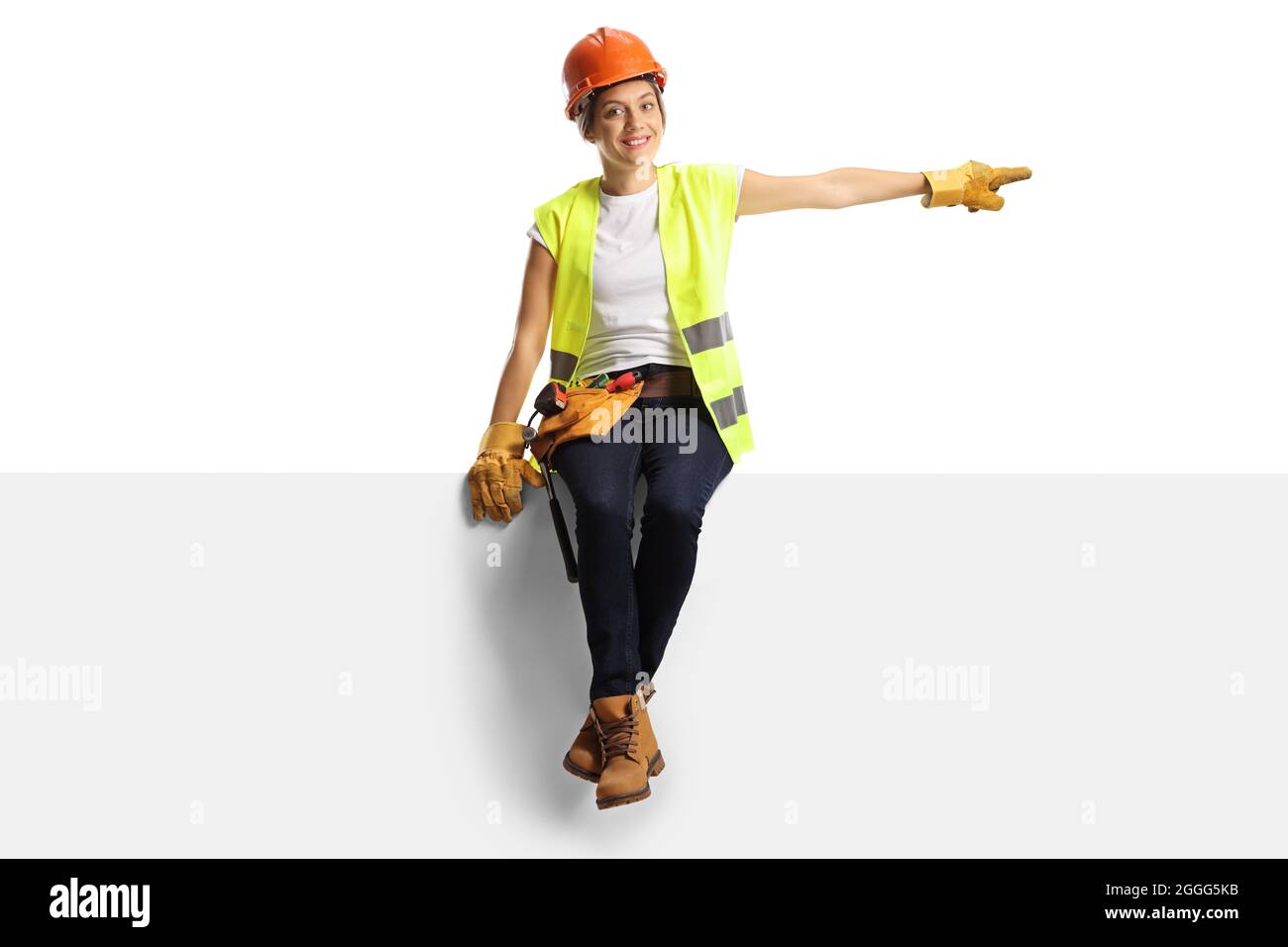 Femme de construction portant un casque et une ceinture à outils sur un panneau et pointant isolée sur un fond blanc Banque D'Images