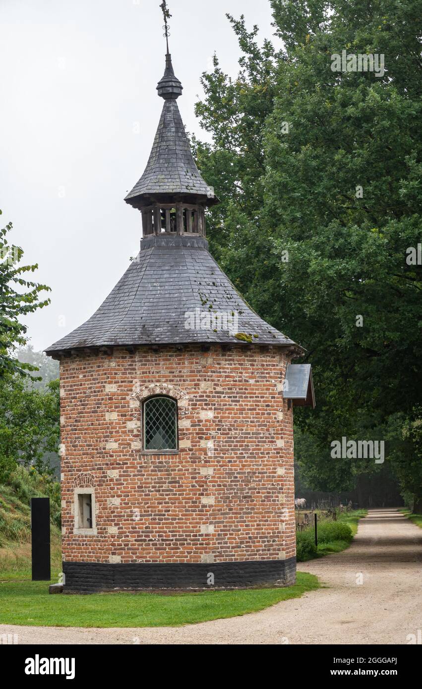 Genk, Belgique - 11 août 2021 : Domein Bokrijk. Brique rouge-rangée chapelle octogonale de Metseren le long de la route de terre sèche avec feuillage vert à l'arrière sous le silv Banque D'Images