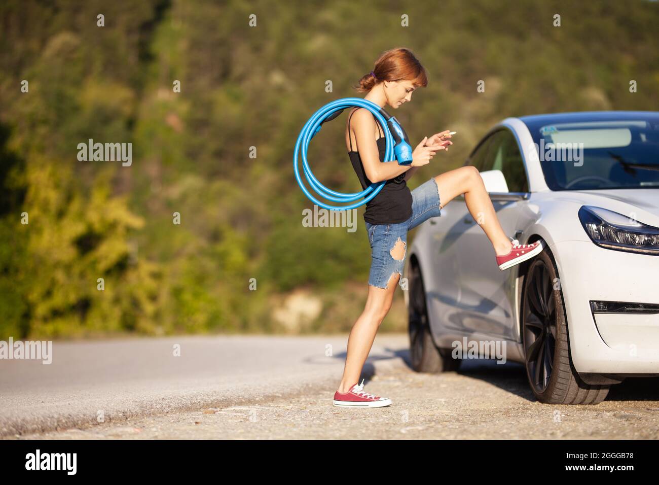 Belle jeune fille à côté d'une voiture électrique. Se tenir debout avec un câble de charge bleu. Banque D'Images