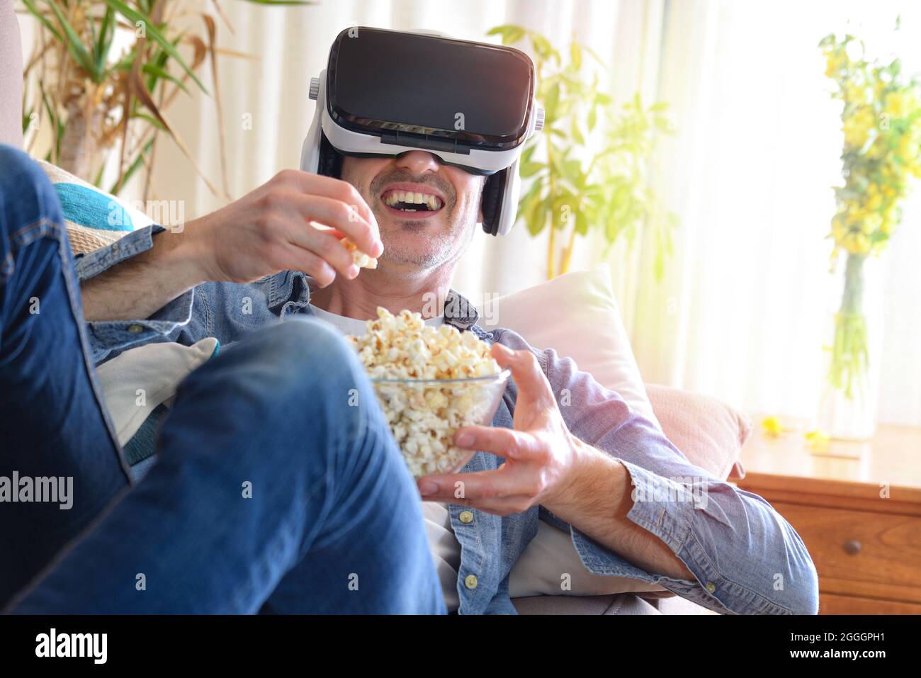 Homme regardant du contenu multimédia dans des lunettes de réalité virtuelle en train de manger du pop-corn allongé sur le canapé dans les détails de la maison Banque D'Images