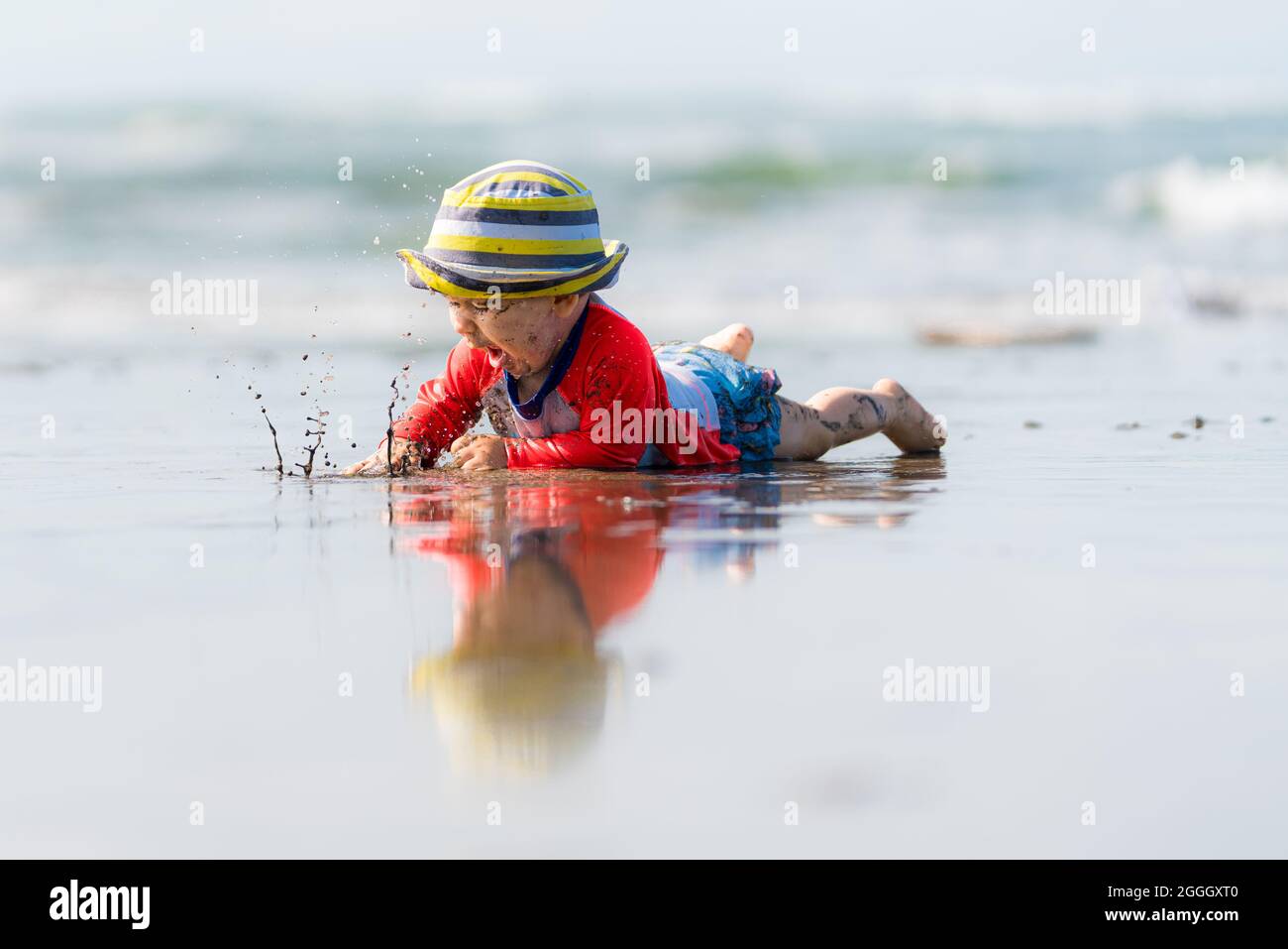 Très beau bébé barbotant de l'eau et rampant à la plage.L'enfant porte un chapeau rayé.Sa réflexion peut être vue sur l'eau. Banque D'Images