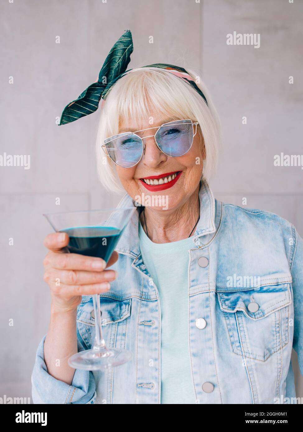 senior (ancienne) femme élégante avec cheveux gris et en verres bleus et  veste en denim tenant verre avec cocktail bleu. Alcool, détente, vacances,  retraite Photo Stock - Alamy