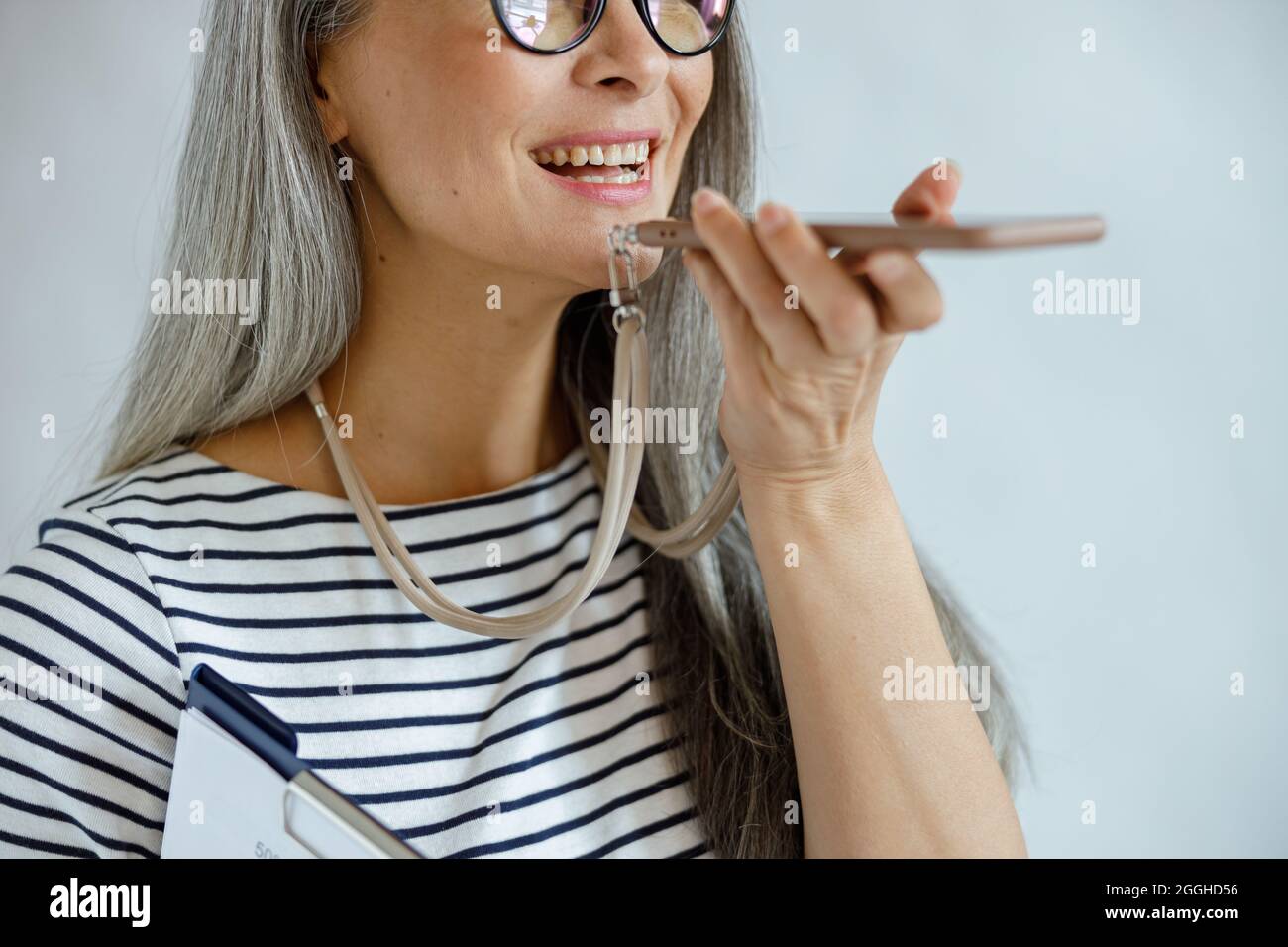 La dame mature positive avec presse-papiers utilise le mode haut-parleur sur le téléphone sur fond gris clair Banque D'Images