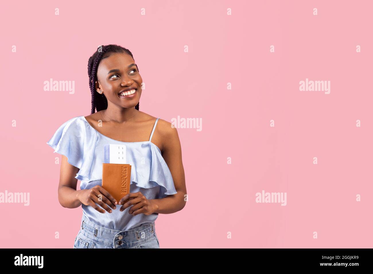 Bonne jeune femme noire avec passeport et billets d'avion en regardant de côté l'espace libre, voyageant à l'étranger sur fond rose Banque D'Images