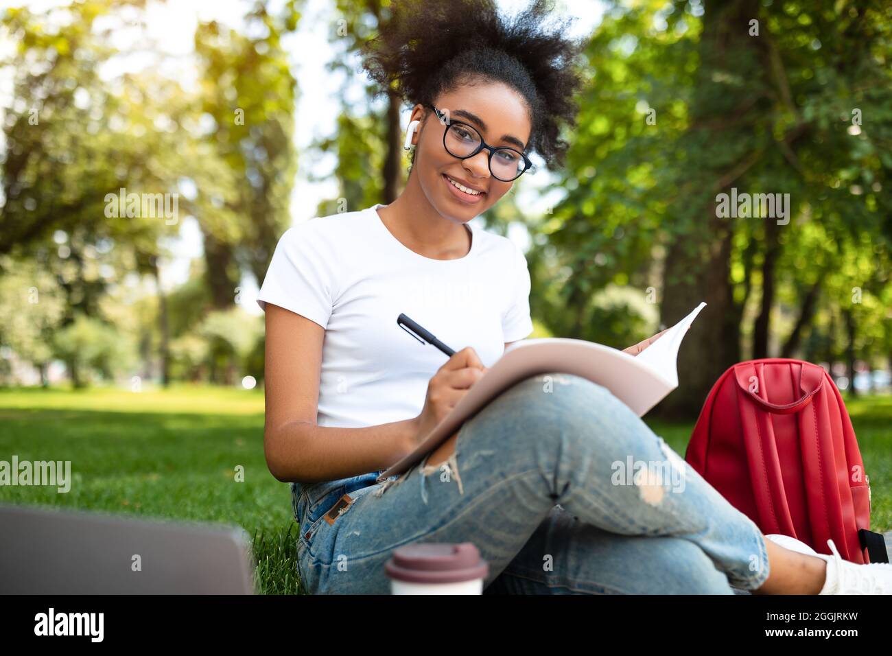Joyeuse Black College Girl prenant des notes d'apprentissage assis dans le parc Banque D'Images