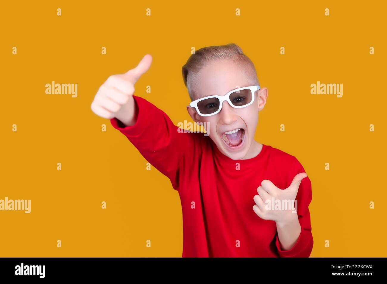 un enfant gai dans des vêtements et des lunettes rouges montre les pouces sur un fond jaune Banque D'Images