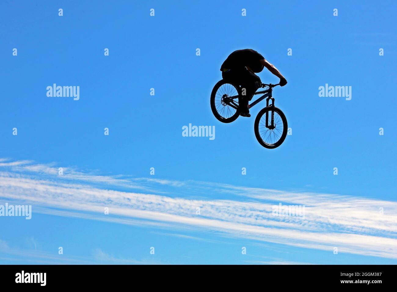 Silhouette de BMX ou de motard de montagne en hauteur lors d'un spectacle de cascades, dans un ciel principalement bleu clair avec une bande de nuages blancs Banque D'Images