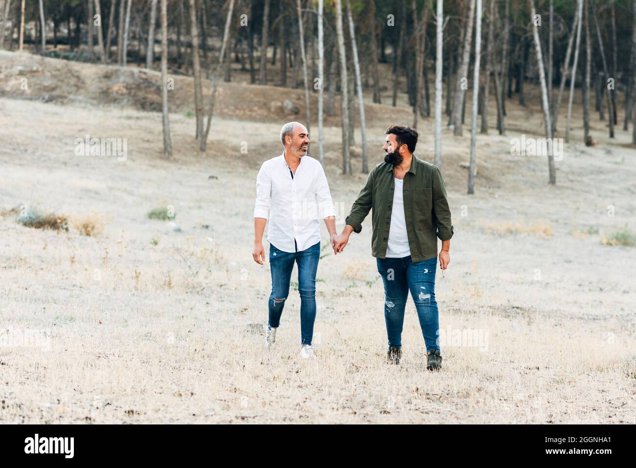 Couple heureux de gays marchant dans la nature tout en tenant les mains. Les garçons lgtb se regardent les uns les autres avec des sourires dans leurs yeux. Banque D'Images