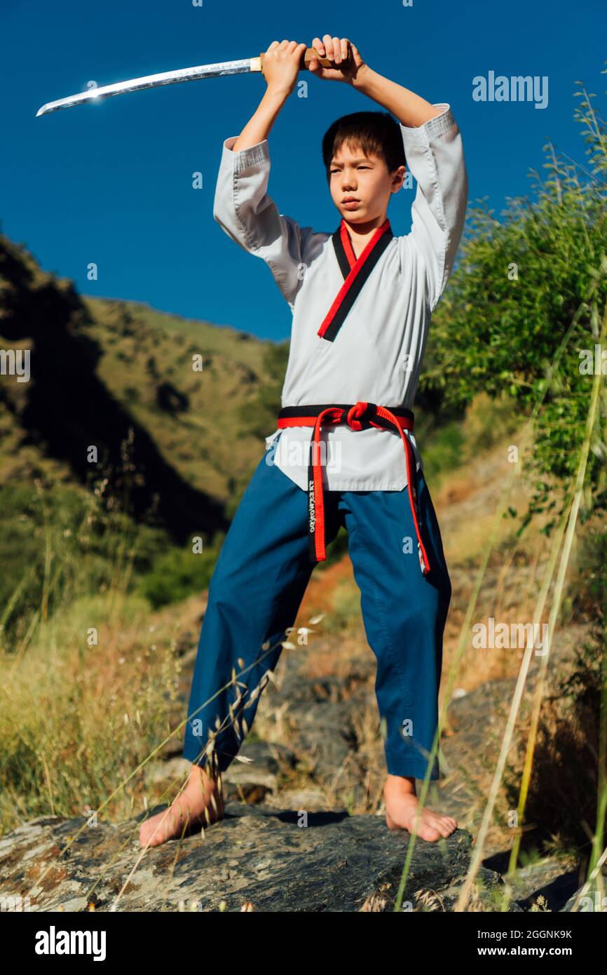 Le garçon ninja japonais lève l'épée de taekwondo en position d'attaque  avec ses mains.il porte un kimono de taekwondo de régulation et une ceinture  noire. Il est au niveau Photo Stock -