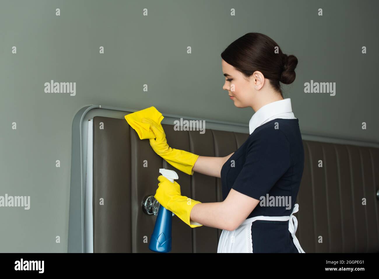 vue latérale de la jeune femme de ménage dans des gants en caoutchouc tenant une bouteille vaporisatrice et un chiffon pendant le nettoyage de la chambre d'hôtel Banque D'Images
