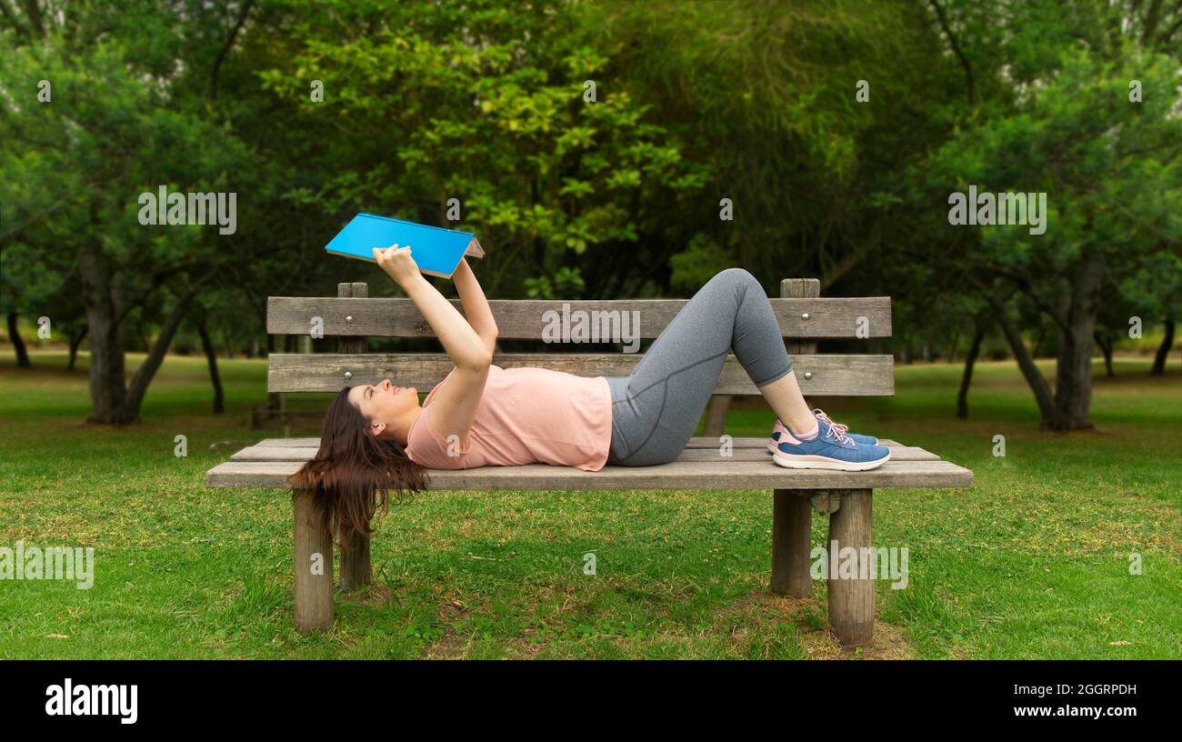 Belle jeune femme hispanique dans les vêtements de sport couché sur le banc en bois au milieu d'un parc sans que les gens lisent un livre bleu sur un fond Banque D'Images