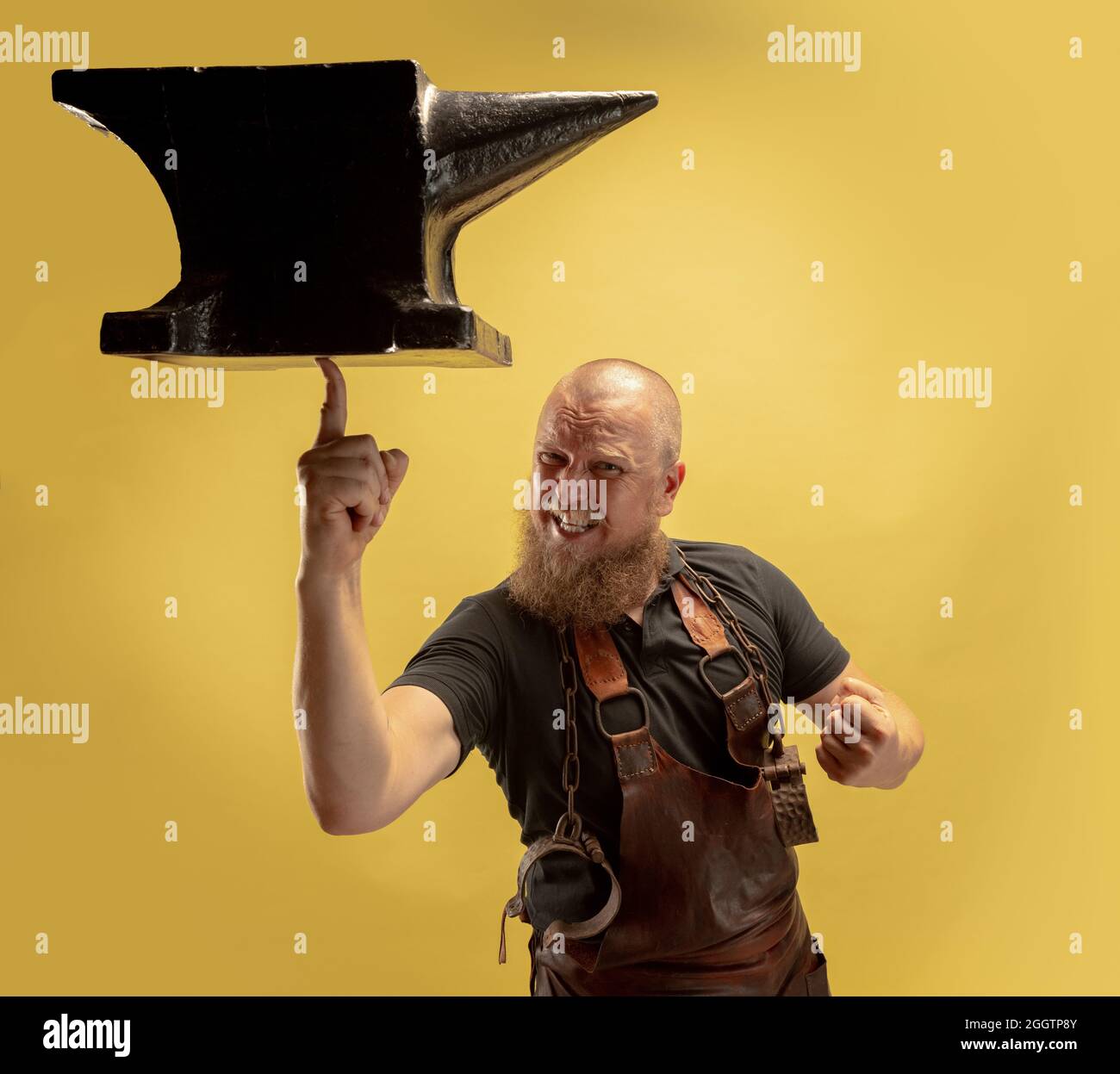 Portrait comique de chauve barbu musclé, forgeron en tablier de cuir ou uniforme isolé sur fond jaune de studio. Concept de travail, rétro Banque D'Images