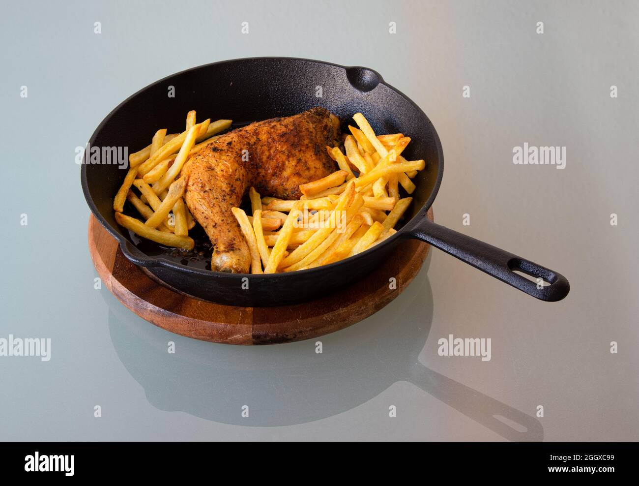 Vue en grand angle de la cuisse de poulet rôtie et des frites dans une poêle en fonte sur une assiette en bois. Réflexion sur table en verre Banque D'Images