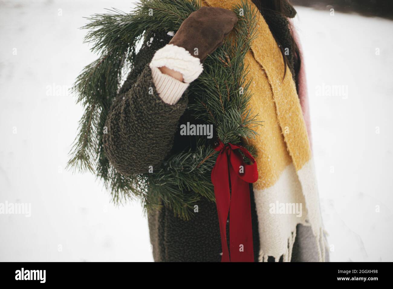Belle femme élégante avec couronne de noël dans un champ d'hiver enneigé.  Jeune femme en moufles confortables et manteau tenant une couronne de Noël  avec noeud rouge. Vacances Photo Stock - Alamy