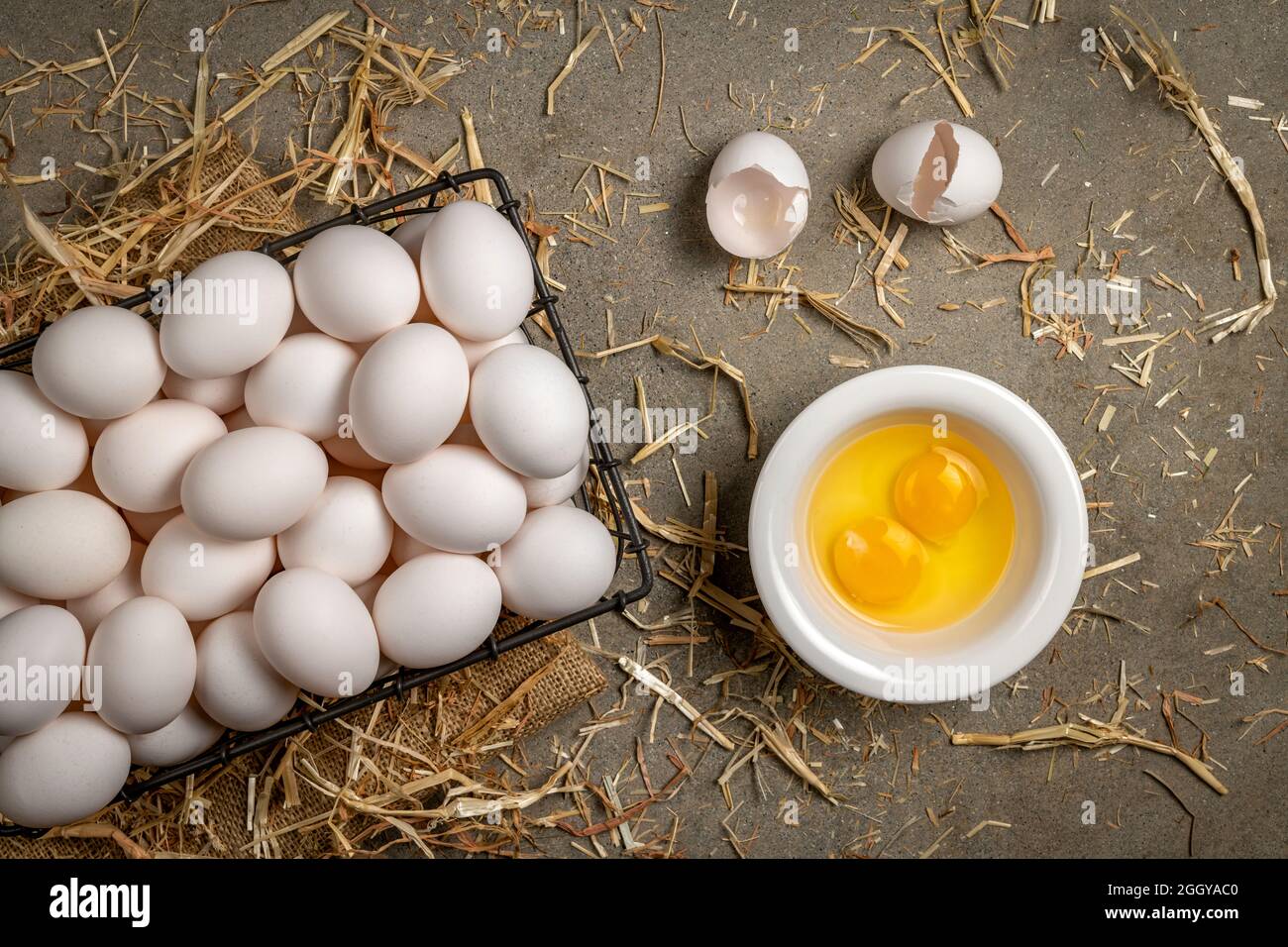 Un petit panier en fil métallique d'œufs frais avec deux œufs fissurés et jaune repose sur un plan d'examen en ciment avec du foin pendant la préparation du petit déjeuner. Banque D'Images