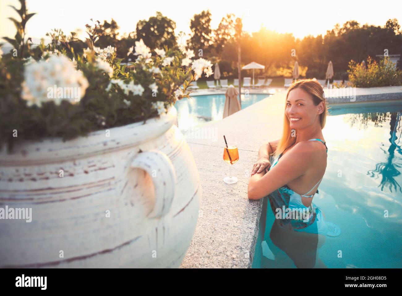 Belle femme souriante dans la piscine avec un cocktail. Lumière chaude et soleil intense en soirée. Banque D'Images