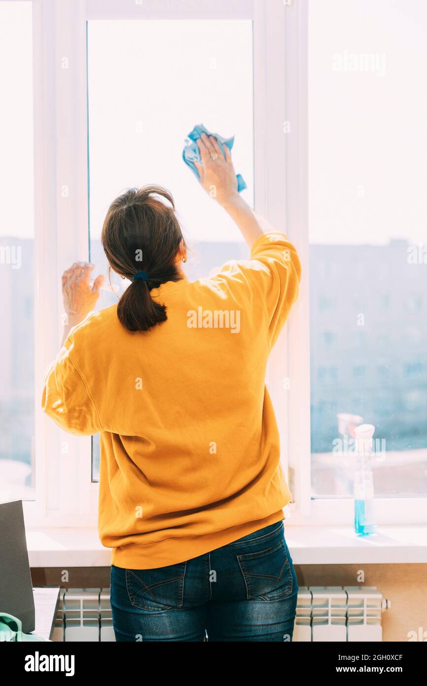 Femme de cinquante en chandail jaune et jeans lave fenêtre poussiéreuse  dans l'appartement. Une femme de 50 ans nettoie les fenêtres des taches à  l'aide de Rag and Spray Photo Stock -