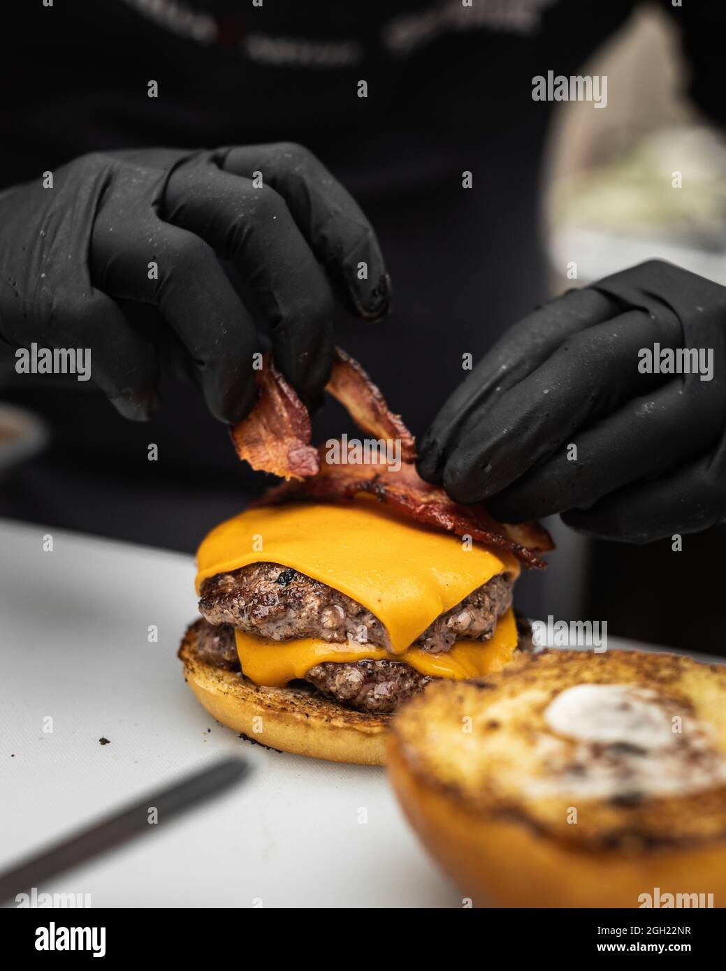 Gros plan d'un cuisinier plaçant le bacon frit sur une tranche de fromage tout en superposant un hamburger Banque D'Images