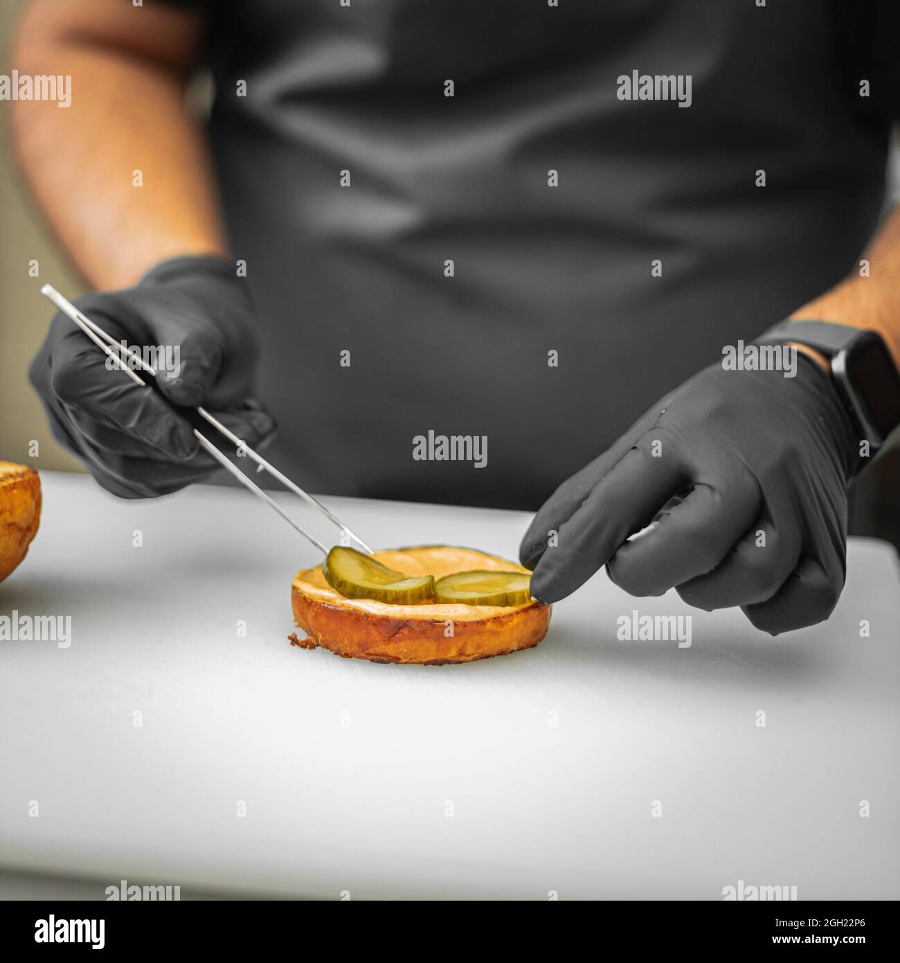 Gros plan d'un cuisinier plaçant des cornichons en tranches sur la sauce tout en superposant un hamburger Banque D'Images