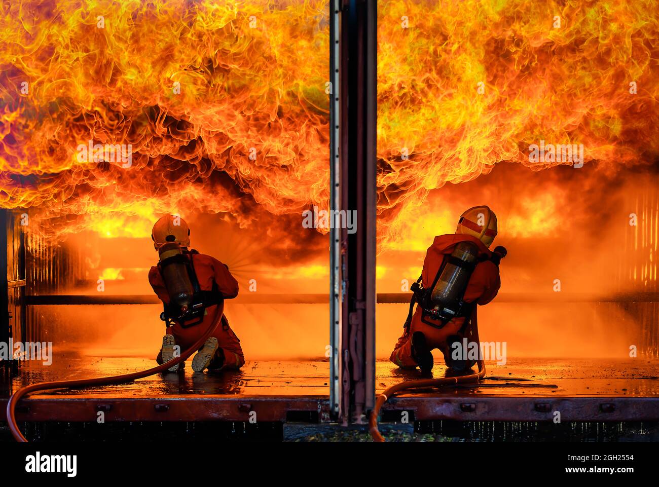 Une équipe de deux pompiers portant un uniforme complet avec réservoir d'oxygène à l'arrière tenant l'eau de propagation de pipe et face à un grand feu d'orange chaud avec brave et Banque D'Images