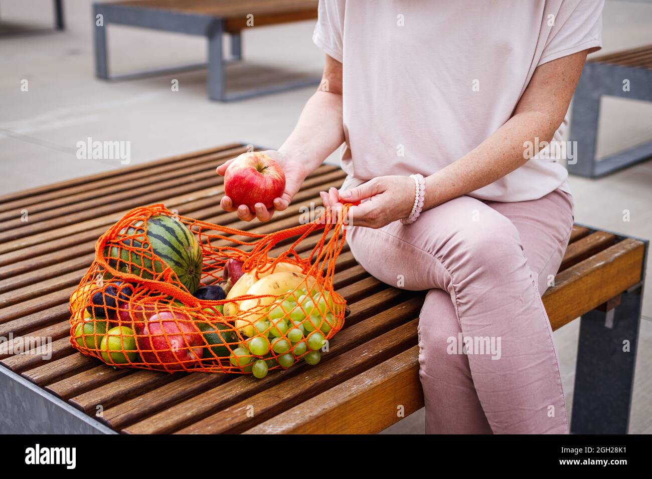 Femme avec sac en maille réutilisable assise sur un banc en ville. Se reposer au banc après avoir fait des achats de fruits au supermarché. Concept zéro déchet et sans plastique Banque D'Images
