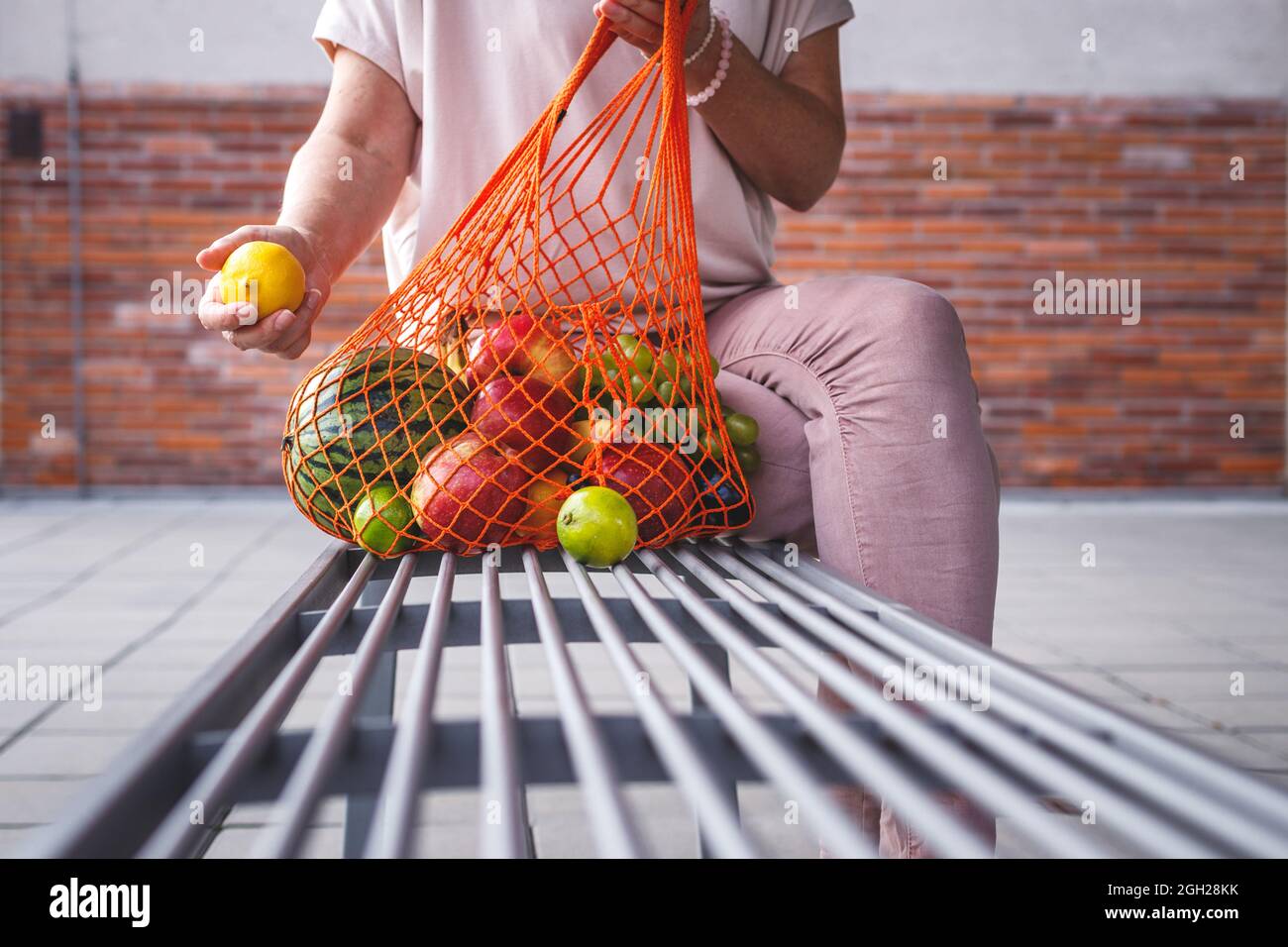 Femme avec sac en maille réutilisable assise sur un banc en ville. Se reposer au banc après avoir fait des achats de fruits au supermarché. Concept zéro déchet et sans plastique Banque D'Images