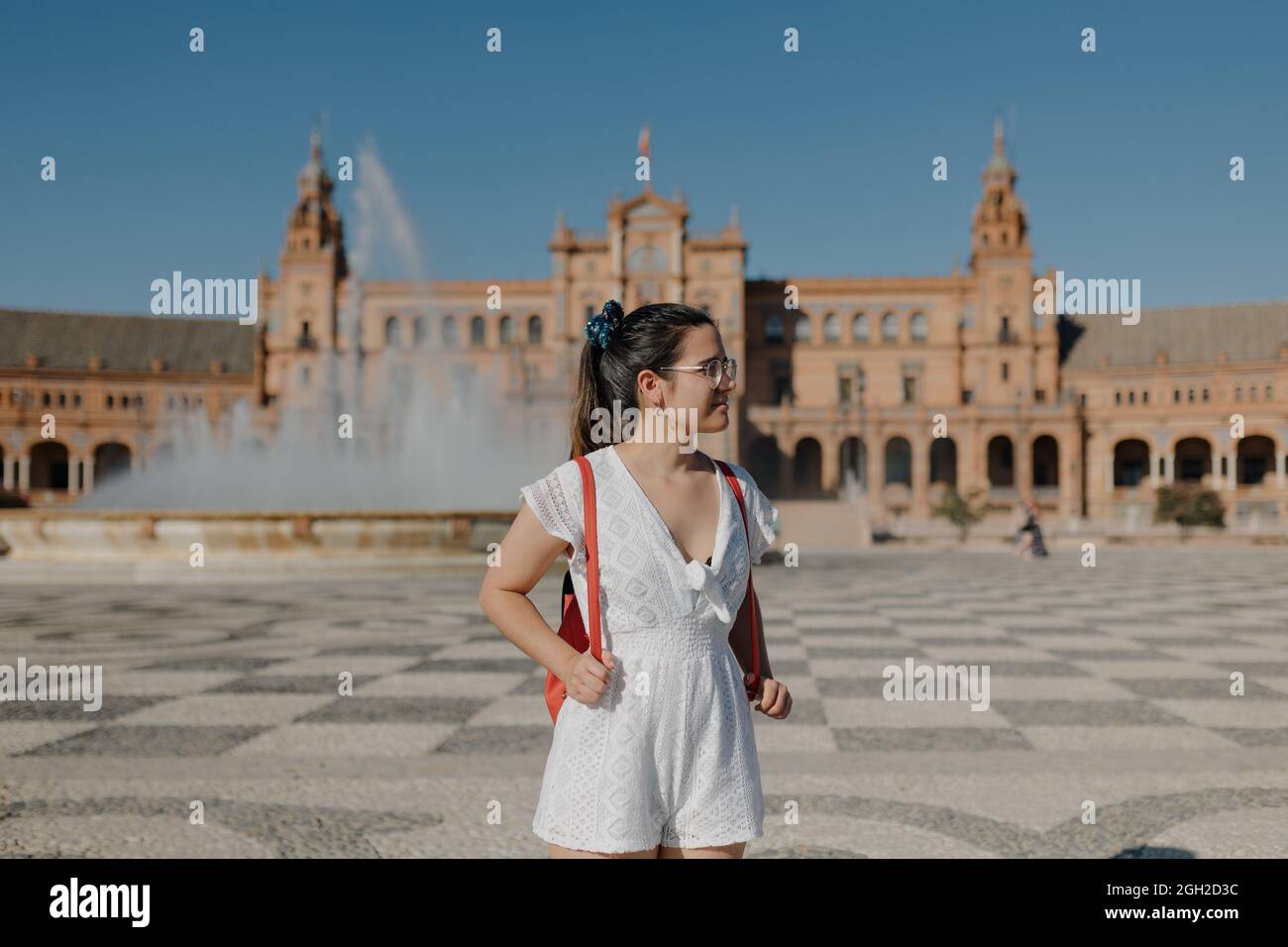 Une jeune femme touristique avec des lunettes portant une robe blanche et un sac à dos rouge regarde loin et sourit tout en se tenant sur la Plaza de España de Séville. Banque D'Images