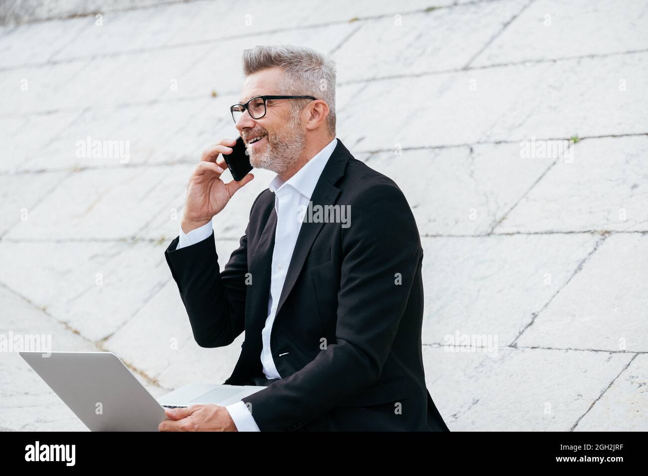 Beau jeune homme d'affaires assis sur les escaliers à l'extérieur travaillant avec son ordinateur portable tout en parlant sur son téléphone. Travail à distance Banque D'Images