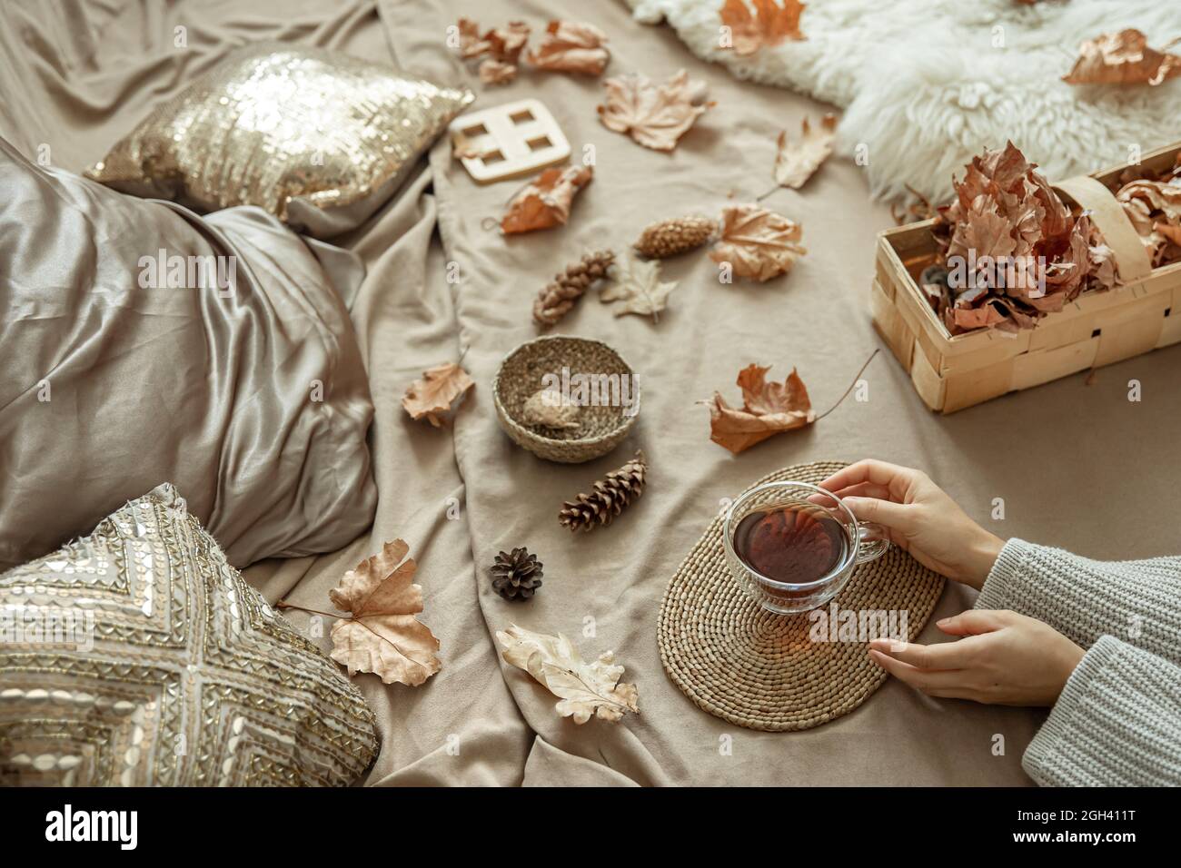 Accueil composition automnale avec les mains des femmes et une tasse de thé au lit, parmi les cônes de pin et les feuilles d'automne. Banque D'Images