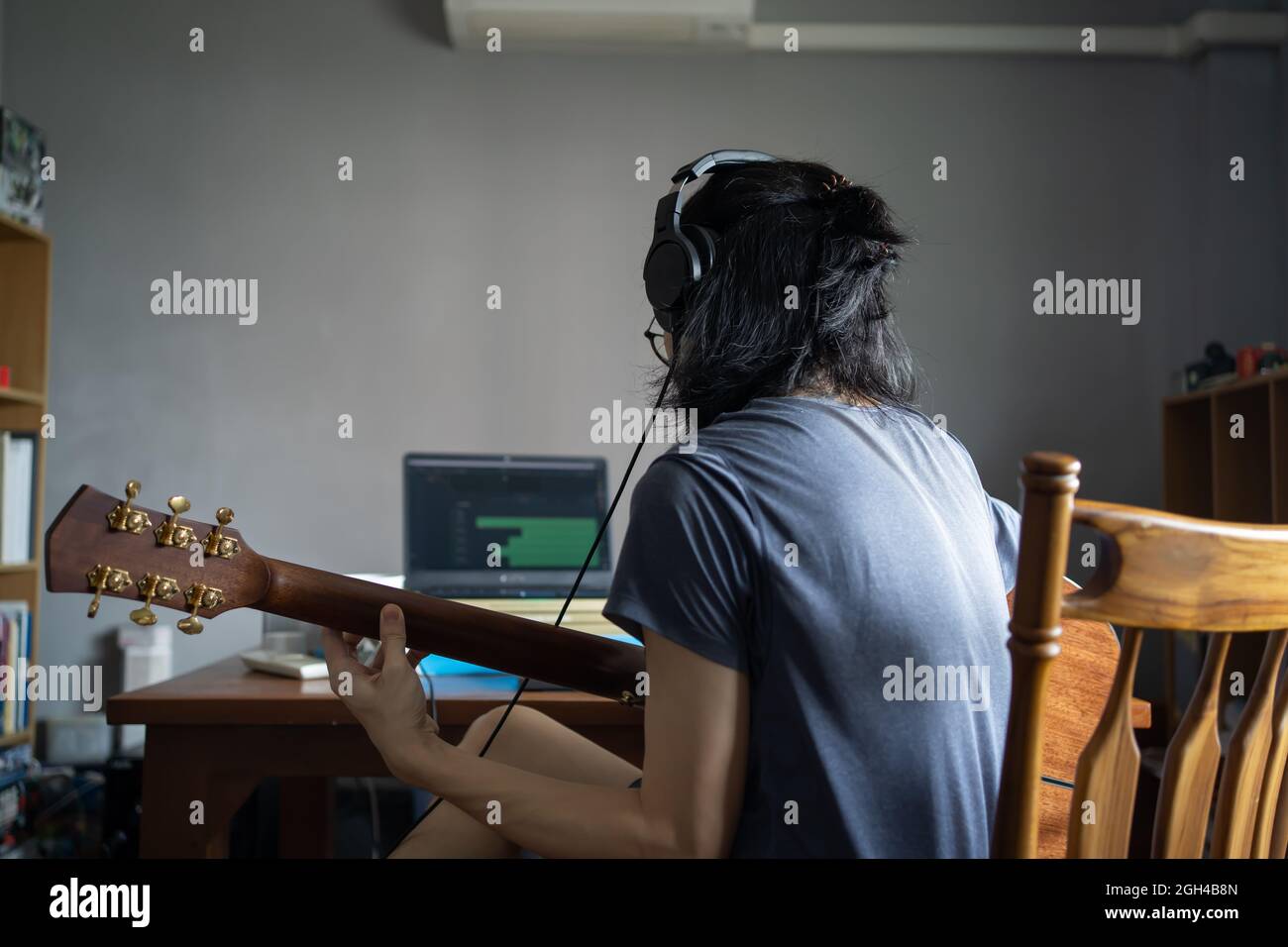 Un homme aux cheveux longs portant des écouteurs et jouant de la guitare acoustique afin d'enregistrer dans un ordinateur dans une pièce domestique Banque D'Images