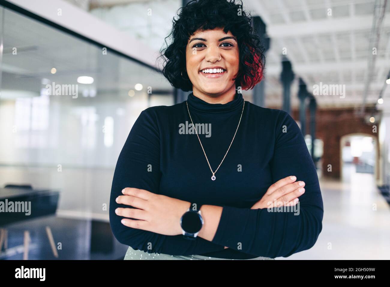 Femme entrepreneur souriant dans un bureau créatif. Bonne femme d'affaires de taille moyenne qui regarde la caméra en se tenant debout avec ses bras croisés. Réussi Banque D'Images Femme entrepreneur souriant dans un bureau créatif. Bonne femme d'affaires de taille moyenne qui regarde la caméra en se tenant debout avec ses bras croisés. Réussi Banque D'Images