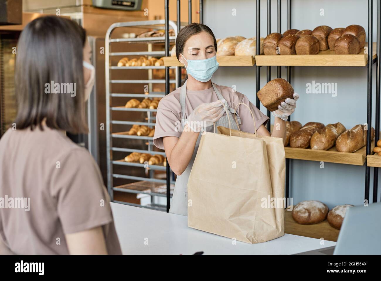 Un jeune employé de boulangerie en tablier met du pain frais dans un sac de papier tout en servant les clients Banque D'Images