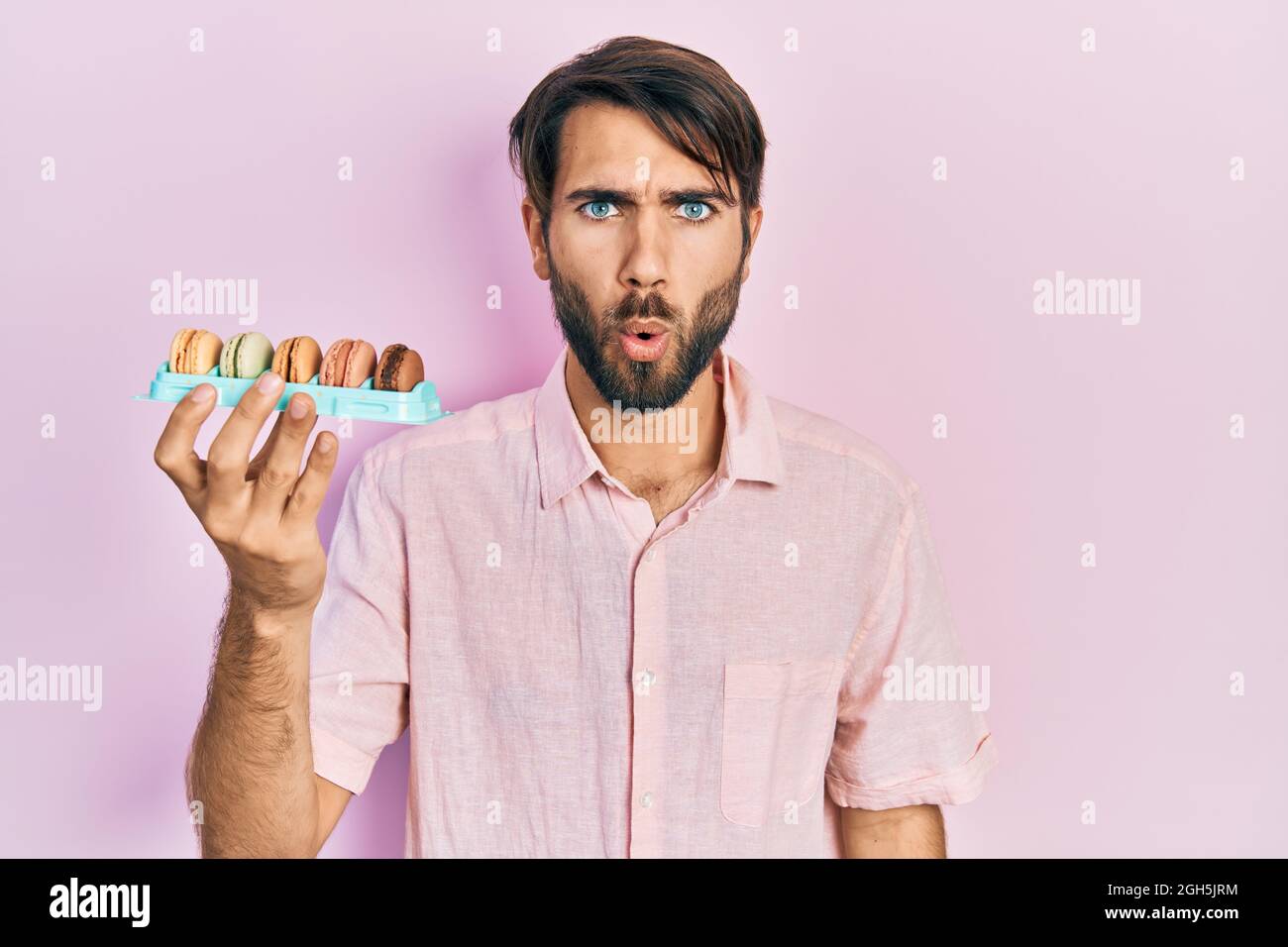 Jeune homme hispanique tenant de délicieuses pâtisseries de macrons effrayés et étonnés par la bouche ouverte pour la surprise, le visage incrédulé Banque D'Images