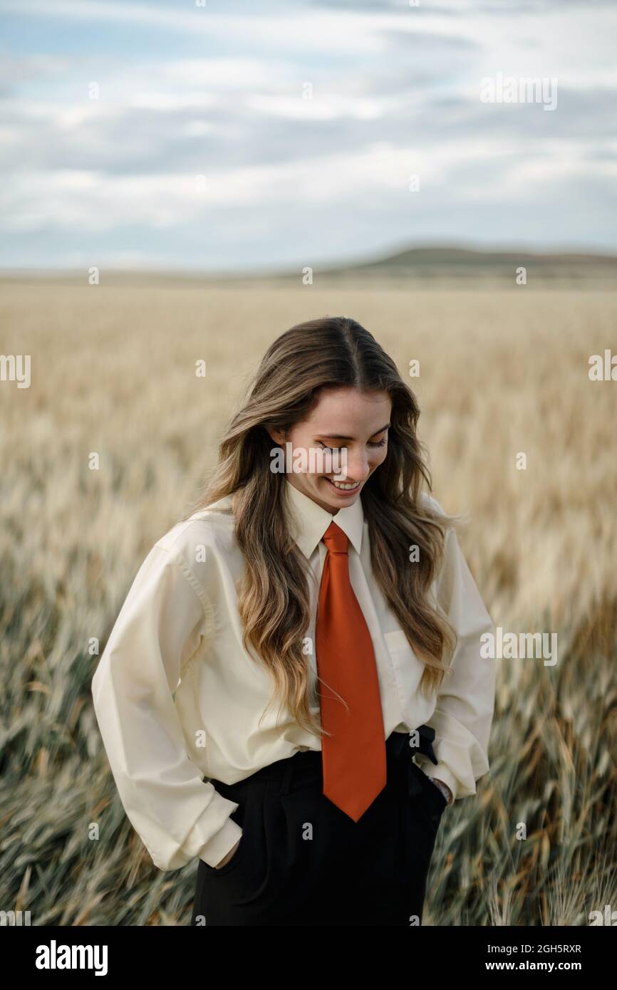 Jeune femme positive en chemise blanche et cravate rouge debout avec les  mains derrière le dos parmi les épis de blé en campagne Photo Stock - Alamy