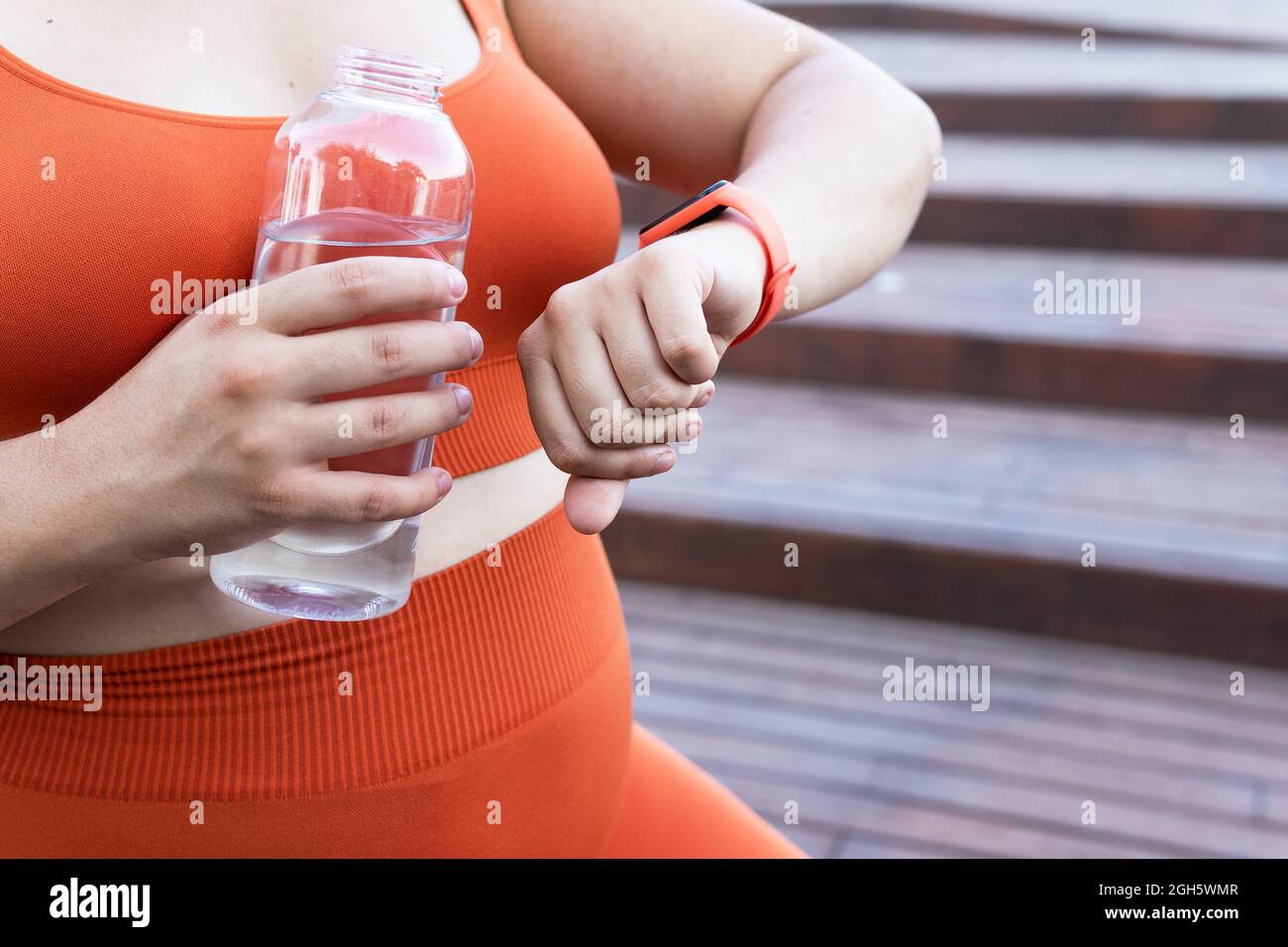 Croise une athlète féminine de taille plus méconnaissable avec une bouteille d'aqua observant la fréquence cardiaque sur un tracker portable pendant l'entraînement dans les escaliers urbains Banque D'Images