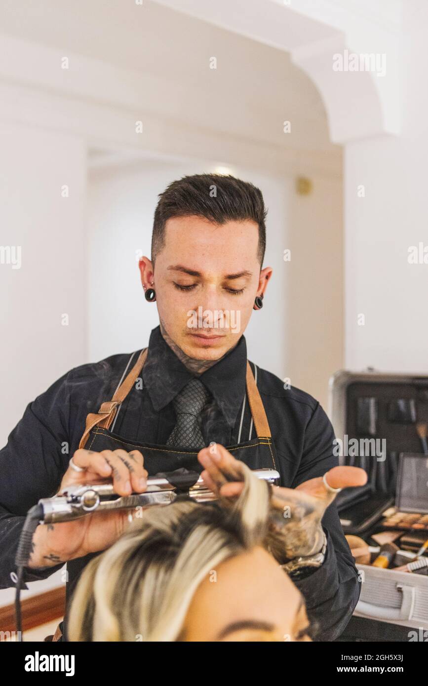Coiffeur masculin utilisant le fer à repasser pour boucler les boucles blondes de la cliente pendant le travail dans le salon de beauté Banque D'Images