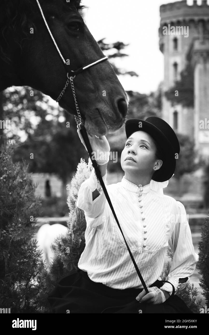 Femme noire adulte sérieuse en chapeau et tenue élégante avec gants pétting cheval brun près des plantes vertes et des arbres dans la campagne Banque D'Images