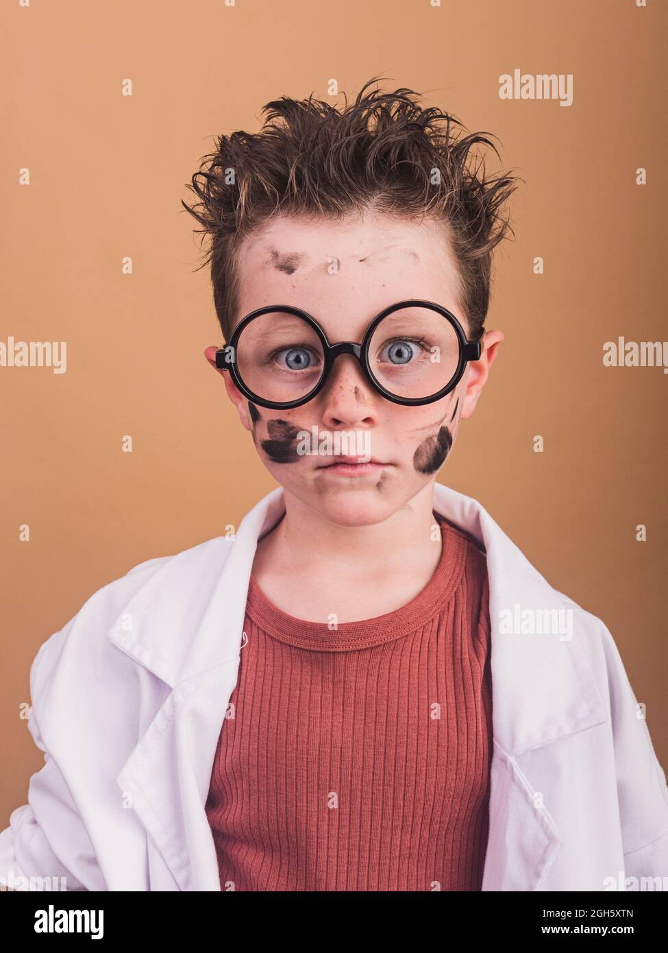 Chimiste garçon dans un peignoir de laboratoire et des verres en plastique regardant l'appareil photo avec un regard fou sur fond beige Banque D'Images
