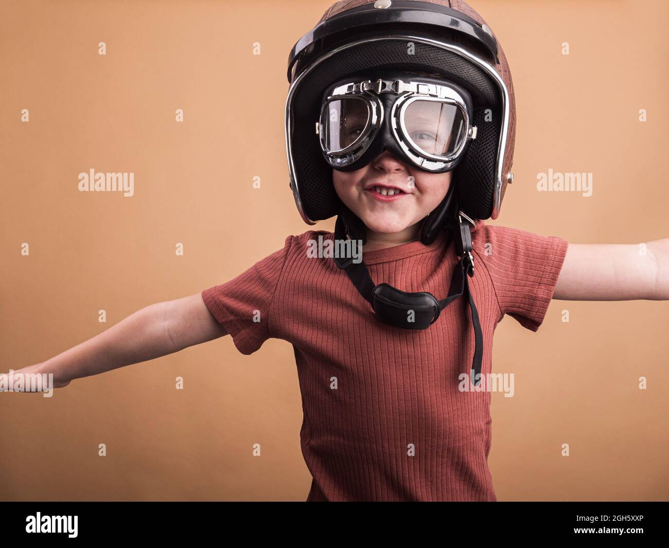 Enfant gai dans un casque et des lunettes de protection regardant la caméra avec des bras étirés sur fond beige Banque D'Images
