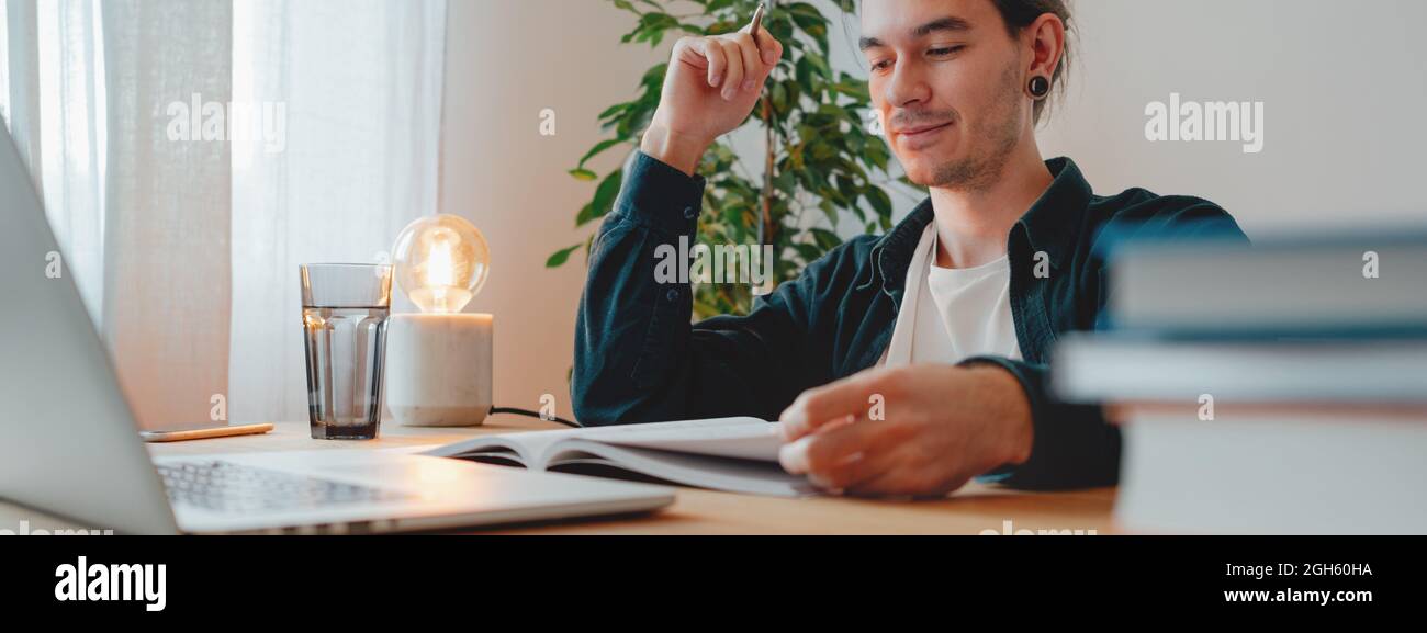 Étudiant à l'université tout en étudiant l'apprentissage virtuel pour passer l'examen à l'université. Un jeune homme étudie à la maison avec un ordinateur portable. Image large Banque D'Images