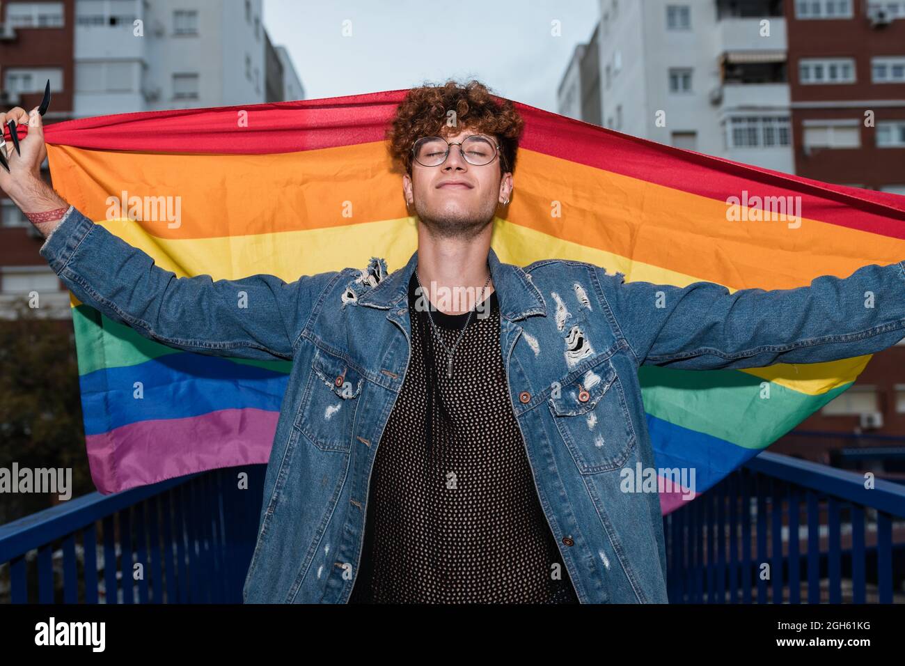 De dessous de l'homosexuel heureux mâle debout avec le drapeau arc-en-ciel dans les bras levés sur le pont et les yeux fermés Banque D'Images