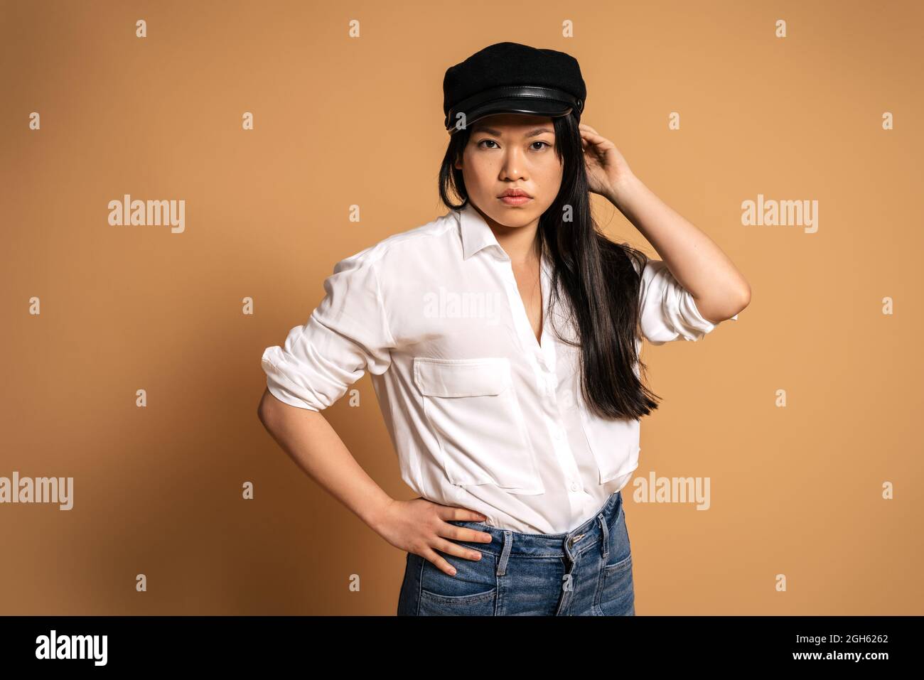 Vue avant du modèle asiatique féminin tendance en chemise blanche et Jean touchant la casquette sur fond beige et regardant l'appareil photo Banque D'Images