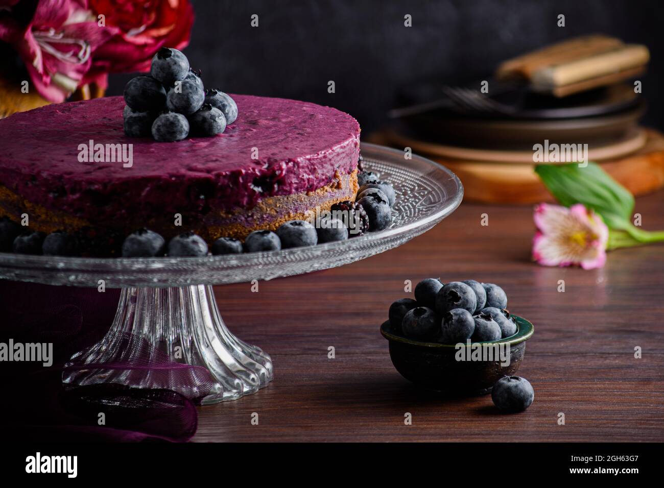 Délicieux gâteau à la mousse aux myrtilles avec crème pourpre décorée de baies fraîches servi sur un support en verre sur table sombre avec fleurs Banque D'Images